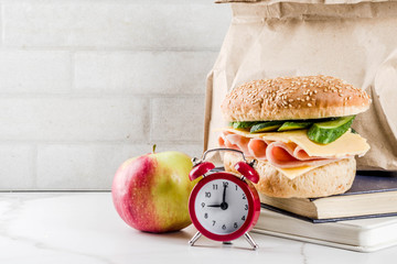 Healthy school food concept, paper bag with lunch, apple, sandwich, books and alarm clock on white kitchen table copy space