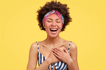 Joyful African American female youngster expresses happines, laughs pleasantly, keeps hands on heart, wears casual clothes, closes eyes in pleasure, stands against yellow background. Emotions concept