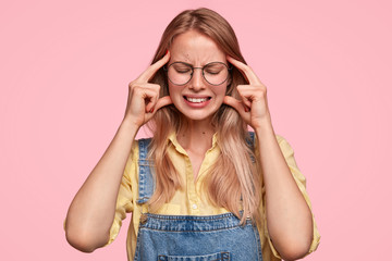 Depressed beautiful female feels stressed after long hours work, touches temples, clenches teeth and suffers from headache, dressed in denim dungarees, stands against pink wall. Tiredness concept