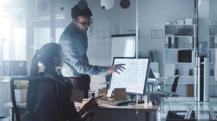 Late at Night/Early Morning in the Office, Male and Female Engineers Discuss  Blueprints Shown on Her Display. Office Looks Modern, with Blueprints on Walls.