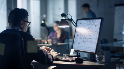 Late at Night in the Office. Design Engineer Works on His Personal Computer. On His Display We See Blueprints. Office Looks Modern. In the Background People Working.