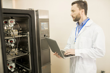 Wall Mural - Portrait shot of highly professional engineer using modern laptop while standing at control cabinet and adjusting pressure transducers