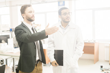 Wall Mural - Bearded middle-aged businessman giving tour of production department of modern factory, young Arabian inspector wearing lab coat listening to him with interest