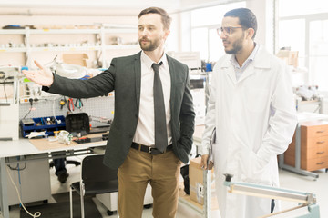 Wall Mural - Handsome middle-aged entrepreneur in formalwear discussing working process with lab technician while standing at spacious production department of manometer factory