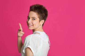 beautiful young girl with short hairdo in a white t-shirt on a pink background pointing a finger to the side