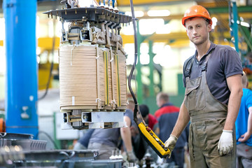 Industrial man worker in factory workshop with conveyor during power transformer assembling by gantry crane