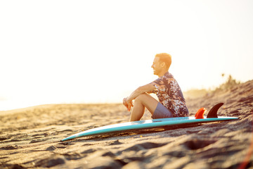 Wall Mural - Portrait of happy surfer in hawaiian t-shirt walking with surf board on the beach at sunset