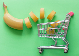 Wall Mural - Yellow cut banana with supermarket cart on green background. Natural light