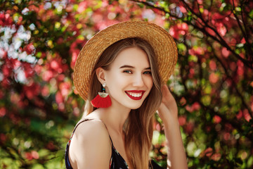 Outdoor portrait of young beautiful happy smiling girl with red lips, long hair, wearing straw hat, long tassel earrings, cold shoulder dress, posing naer blooming tree. Copy, empty space for text