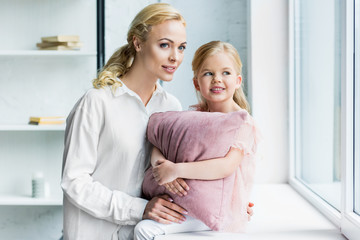 mother and adorable smiling daughter with pillow looking at window at home