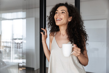 Sticker - Photo of young curly woman with long dark hair smiling and looking aside, while standing near glass door in house with cup of tea