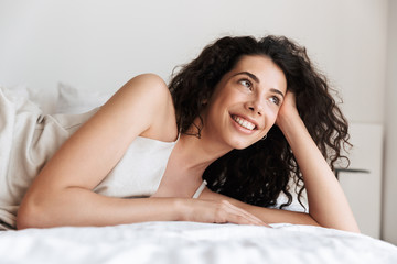 Sticker - Closeup photo of pretty young woman 20s with long curly hair wearing silk leisure clothing lying in hotel bed, and looking aside while propping up her head with hand