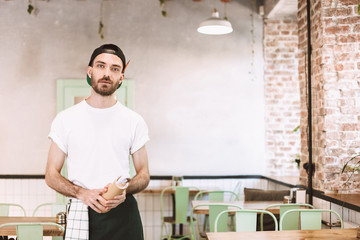 Young waiter in black cap,white t-shirt and apron standing with notepad in hands and dreamily looking in camera while working in cafe