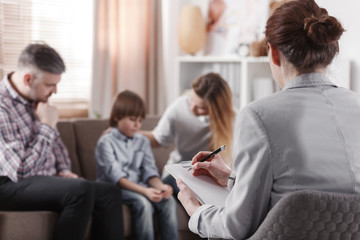 Wall Mural - Psychologist taking notes while listening to a family with a boy sitting on a sofa in the office