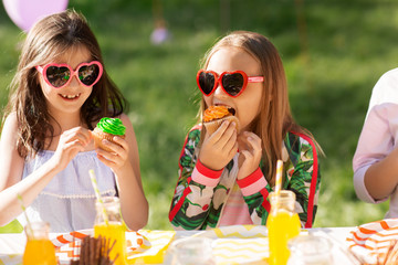 Poster - holidays, childhood and celebration concept - happy kids in sunglasses sitting at table on birthday party at summer garden and eating cupcakes