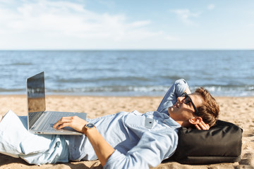 Young guy with glasses, lying on the sand, working on his laptop on the beach, against the sea, working on vacation, suitable for advertising, text insertion