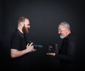 Two male photographers, young and elderly, with cameras - modern digital and vintage widescreen. On a black background in the studio.