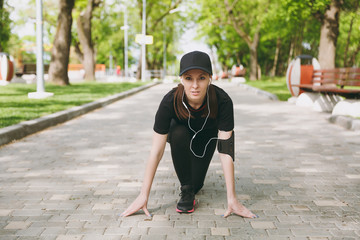 Young athletic beautiful brunette girl in black uniform and cap with headphones listening music at low start before running, training on path in city park outdoors. Fitness, healthy lifestyle concept.