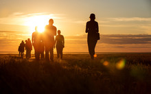 A Group Of People Walking In A Track. They Go Against The Background Of The Orange Sun, Their Contours And Silhouettes Are Visible.
