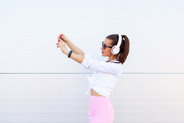 Wall Mural - Fashion portrait of pretty smiling woman in sunglasses making selfie by the smartphone against the grey wall. Wireless Headphones, fitness bracelet on the hand