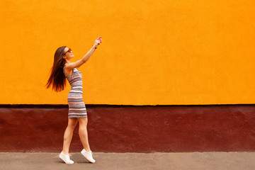 Wall Mural - Fashion portrait of pretty smiling woman in sunglasses making selfie by the smartphone against the orange wall.Fitness bracelet on the hand. flying hairs