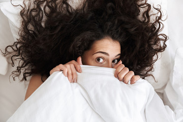 Wall Mural - Portrait from above of cute woman 20s with dark curly hair covering her face with white blanket, while lying in bed after waking up in morning