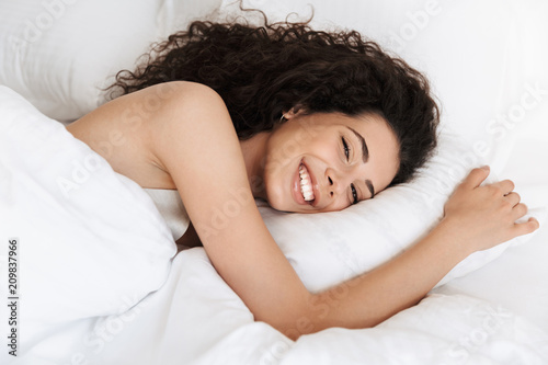 Fotomural Image of beautiful woman 20s with dark curly hair smiling, while lying in bed on