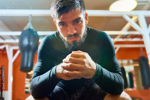 Fotografia, Obraz Confident bearded man in sportswear sitting with hands together and looking at camera on background of boxing gym interior