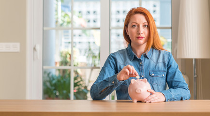 Sticker - Redhead woman saves money in piggy bank at home with a confident expression on smart face thinking serious