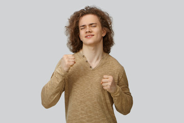 Wall Mural - Portrait of handsome positive young male with wavy hair dancing in studio, keeping eyes closed. Attractive emotional smiling guy clenching fists, expressing true joy, being happy with good news