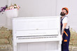 Loader moves piano instrument. Man with beard, worker in overalls and helmet lifts up piano, white background. Delivery service concept. Courier delivers furniture in case of move out, relocation.
