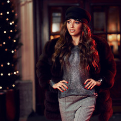 Wall Mural - Fashionable woman with brunette hair wearing cap, fur coat, gray jumper and trousers. Female model posing against building entrance door and potted trees decorated with light garland on background.