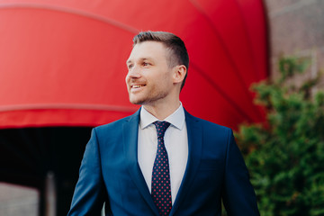 Cheerful manager dressed in formal outfit, has pleasant smile on face, being in good mood after successfully signed contract, poses outdoor against red background. People, success and job concept