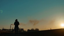 Man Hiker Homeless Person Traveler Walking By Railroad Silhouette Road. Man With A Backpack Going To Sunset Silhouette Against A Background Of An Industrial Zone Plant Pipe Smoke. Man Traveler Concept