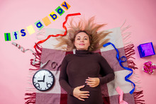 Beautiful And Young Blonde Pregnant Woman Lies On The Floor On A Plaid Next To The Clock And A Cards With The Inscription "it's A Boy" In Studio Pink Background View From Above. Baby Shower Concept