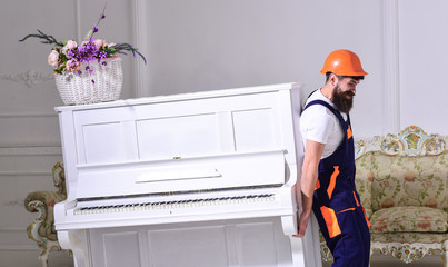 Wall Mural - Loader moves piano instrument. Man with beard, worker in overalls and helmet lifts up piano, white background. Courier delivers furniture in case of move out, relocation. Delivery service concept.