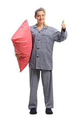 Poster - Young woman in pajamas holding a pillow and making a thumb up sign