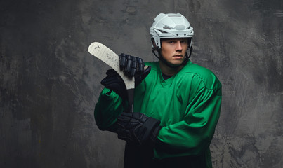 Hockey player wearing green protective gear and white helmet standing with the hockey stick on a gray background.