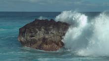 SLOW MOTION: Powerful Ocean Wave Splashes Across A Big Rock In The Middle Of The Rough Exotic Sea. Breathtaking View Of Violent Breaking Wave Foaming Over A Black Rock Near Rocky Tropical Shore.