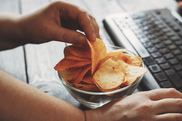 Unhealthy snack at workplace. Hands of woman working at computer and taking chips from the bowl. Bad habits, junk food, high calorie eating, weight gain and lifestyle concept