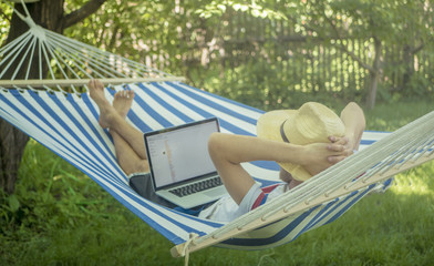 back view of lying young man in hat working with laptop on hammock on a summer day