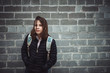 Schoolgirl with backpack stands against gray brick wall with sad emotion in cloudy weather with copy space. Atmospheric portrait of melancholic teenage girl in faded tones. Depressed young girl.