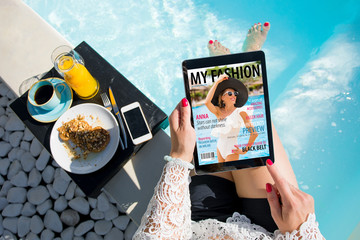 Woman relaxing by the pool and reading emagazine on tablet at breakfast. All contents are made up.