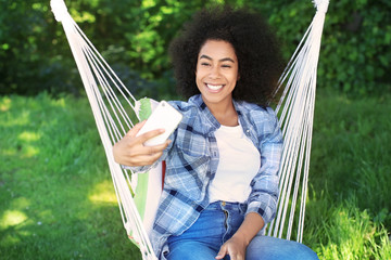 Wall Mural - Beautiful young African-American woman taking selfie in hammock outdoors