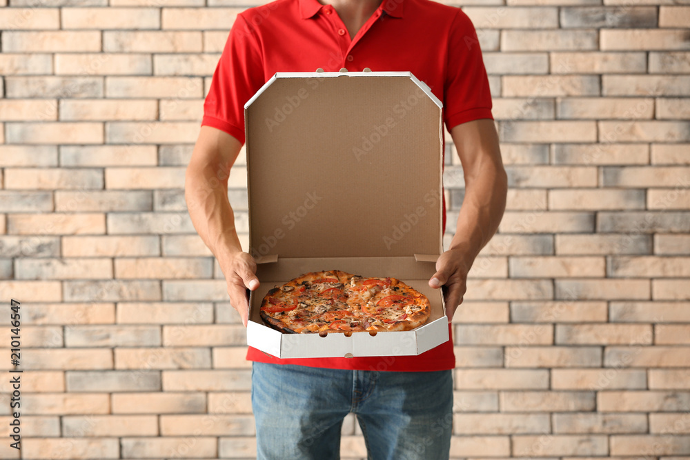 Young man holding box with tasty pizza against brick wall. Food delivery service