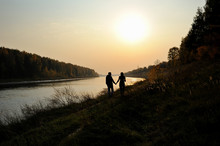 Young Couple Is Walking Along The River Bank At Sunset. Warm Autumn Evening. Silhouettes Of A Girl And A Boy Walking By The Hand. Silhouettes Of Trees On The River Bank And Low Setting Sun In The Sky.