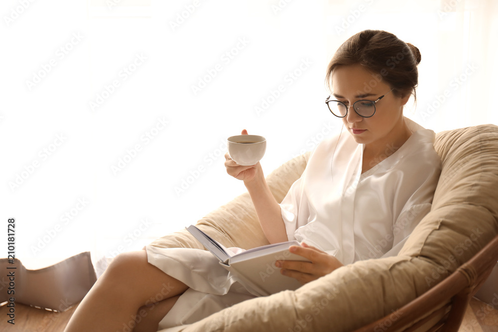 Young woman drinking coffee while reading book on lounge chair at home