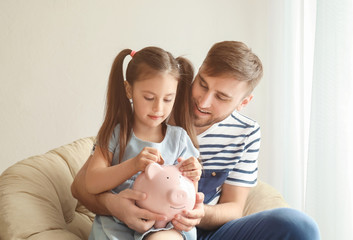 Happy little girl with her father sitting in armchair and putting coin into piggy bank indoors. Money savings concept