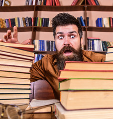 Leinwandbilder - Man on shocked face between piles of books, while studying in library, bookshelves on background. Deadline concept. Teacher or student with beard sits at table with glasses, defocused.