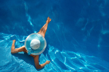 beautiful young woman in wide-brimmed blue hat sunbathing lying in the pool with turquoise clear water